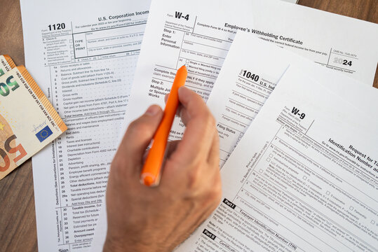 Taxpayer hand points at tax forms with pen learning features and rules of filling blanks avoiding professional assistance services at workplace with euro banknotes