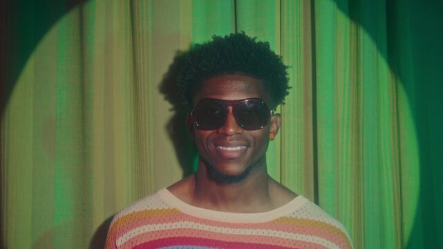 Portrait of young African American man in oversized retro sunglasses and striped knit shirt standing in spotlight and smiling brightly at camera. Zoom-in shot