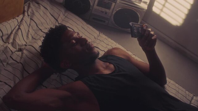 Young Black man lying on bed, holding cassette tape in hand and looking at it thoughtfully while relaxing at home