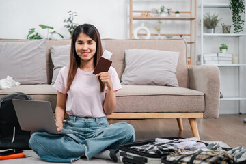 Smiling woman sitting on floor with laptop and passport, ready for travel. Cozy living room with suitcase and casual attire.