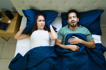 Sleepless woman covering ears with pillow from snoring partner