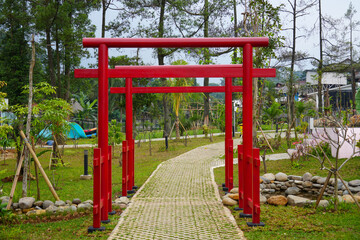 A vibrant red Japanese torii gate standing at the entrance of a garden path, surrounded by lush greenery and tropical trees. The path leads into a tranquil and serene natural setting.