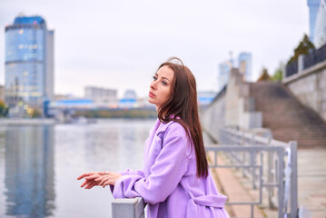 There is a woman in a purple coat standing on the pier, with a view of the pond. She has a beautiful and slender figure.