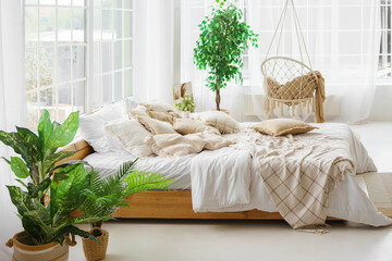 Comfortable bed with lots of soft decorative beige pillows in light sunny bedroom with green plants and large window. Interior design of living room in boho style
