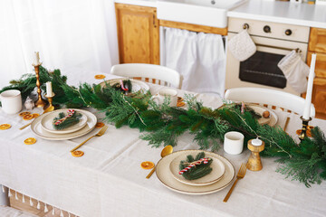 Christmas decor. Beautifully arranged dining table, elegant white plates, golden cutlery and spruce garland on white tablecloth, creating festive holiday atmosphere for meal with family and friends.