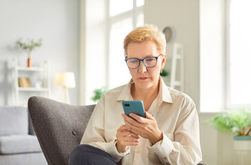 Senior woman in glasses sitting in cozy armchair and using smartphone in living room. Mature lady focusing on screen, browsing or texting, enjoying modern technology in comfortable home environment.