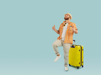 Full body photo of a funny young happy man in beach hat with yellow suitcase isolated on studio blue background. Tourist is going on summer holiday trip. Vacation and travel concept.