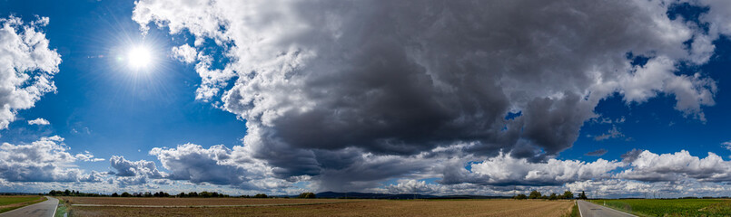 Panoramafoto einer Wolkenlücke mit der Sonne und dunkler Regenwolke im Gegenlicht über...