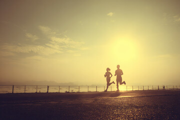 Two people jogging together on paved path near water at sunrise, silhouettes against golden sky, peaceful and energetic morning exercise scene