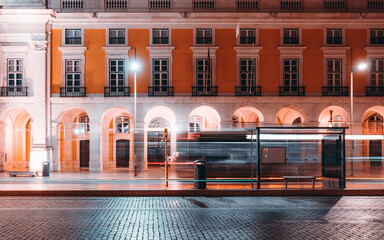 Long exposure night shot of a bus stop in Lisbon with motion blur of passing vehicles, illuminated street lamps, and historic architecture with bright yellow facade in the background © skyNext