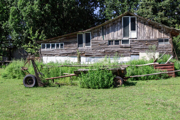 An old wooden barn with windows in an abandoned farm. A metal harrow and horse-drawn cart in front of the barn.