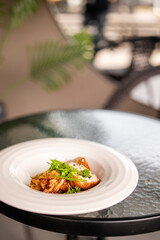 Gourmet meat dish served on a white plate, garnished with herbs and microgreens, presented on a glass table in an outdoor fine dining setting with soft natural light