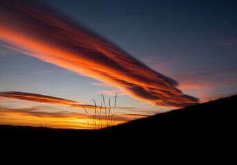 Dramatic lenticular clouds glow orange and red at sunset over a dark hill