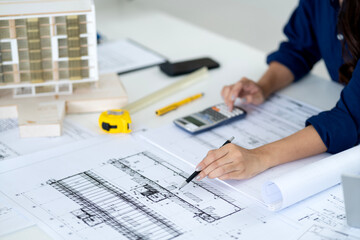 Asian adult architect professional carefully analyzes detailed building blueprints using calculator...