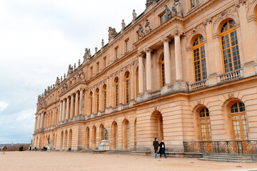 Fototapeta premium Visitors admire the stunning facade of Palatul Versailles, marveling at its intricate architecture and historical significance under a cloudy sky.