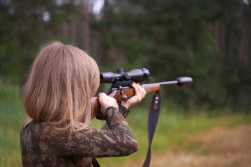 Woman in camouflage aims scoped rifle with concentration, standing in natural surroundings. Hunting in action. Female strength and wilderness survival skills