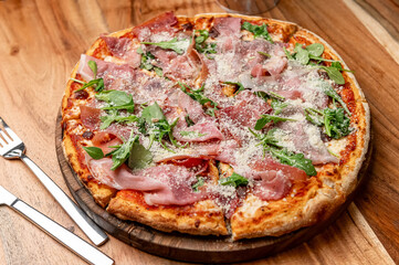 Tasty wood-fired pizza!. A wood-fired pizza topped with prosciutto, arugula, and parmesan cheese is served on a wooden board.