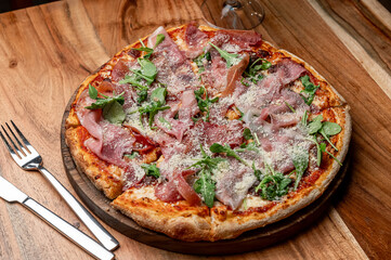 Tasty pizza on wooden board. Thin crust pizza topped with ham, arugula, and cheese served on a wooden board in a cozy dining setting.