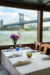 Dining with a view of Manhattan bridge. A table is set for a meal inside a boat, with flowers and city views of Manhattan Bridge in the background.