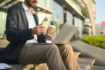 Cropped shot of a White businessman balancing work tools with a laptop, smartphone, and coffee during a busy day in the city