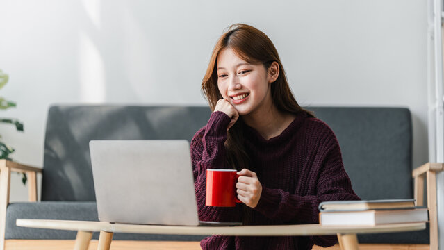 Smiling Asian Woman Student Studying Online with Laptop and Red Mug at Home Happy Young Female Learning Remotely or Watching Movie Enjoying Coffee Break