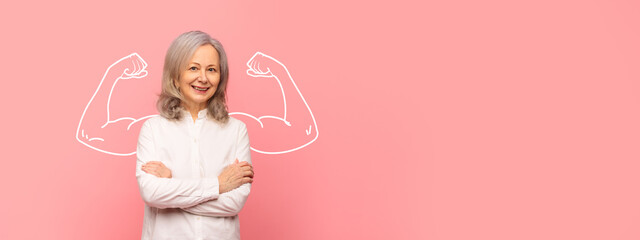 A mature woman stands confidently with her arms crossed, smiling against a bright pink background. Her demeanor reflects strength and empowerment, celebrating resilience and spirit.