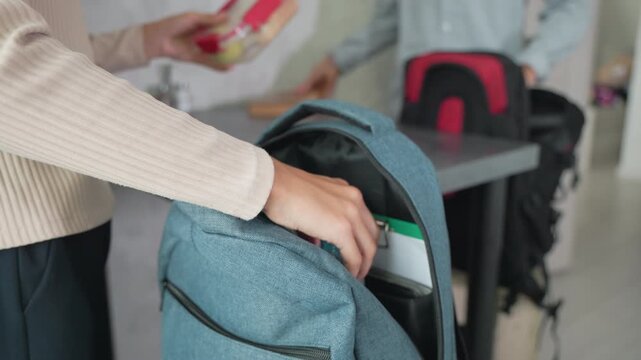 girl and boy in casual wear packing backpacks on kitchen counter placing notebooks water bottles lunch boxes ready for school routine morning preparation with organization and sibling teamwork