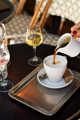Vertical shot of a womans hand pouring coffee into milk foam, with white wine on the table, Paris vibe from woven chairs. Ideal for travel visuals, food scenes, or chic lifestyle with a cozy appeal.