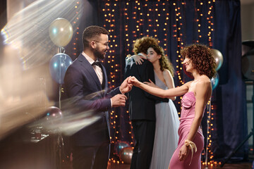 Young adult Caucasian man holding hand of young adult Caucasian woman while dancing at formal event, another young adult couple embracing in background with festive decorations © AnnaStills