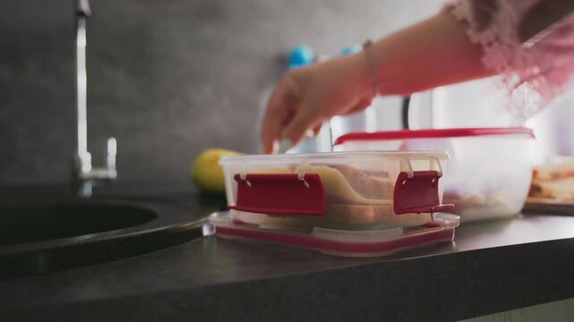 closeup of hand placing sandwich slices into plastic container with red lid on kitchen counter next to sliced bread, green apples, and faucet in bright light preparing healthy school lunch