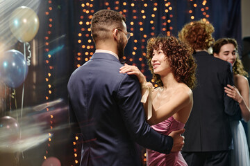 Young adult Caucasian man dancing with young adult Asian woman at formal event, both smiling and making eye contact, other young adults dancing in background, festive atmosphere