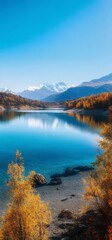 Turquoise Lake Surrounded by Autumn Trees and Distant Snow Capped Mountains Under Clear Blue Sky