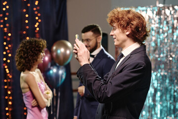Caucasian teenage boy taking photo with smartphone at event, standing in foreground while teenage girl and bearded Caucasian young adult man interacting in background near balloons