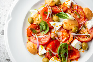 Fresh salad with tomatoes, olives, red onion, basil, white cheese, creamy dressing, and green herb sauce on a white plate. Colorful, appetizing, and healthy food presentation