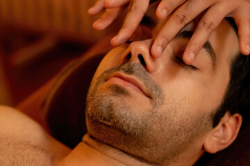 Obraz premium Caucasian man enjoying relaxing anti-stress head massage and pampering facial beauty skin recreation leisure in warm candle lighting ambient salon spa in luxury resort or hotel. Quiescent