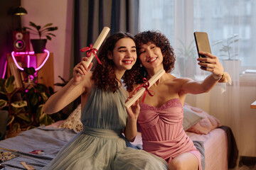 Two Caucasian teenage girls sitting on bed holding diplomas and smiling while taking selfie with smartphone, celebrating graduation together in cozy bedroom setting