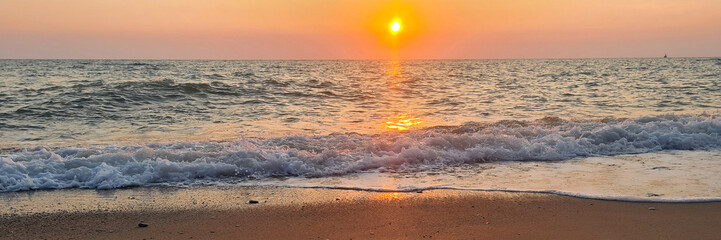 A serene beach scene at sunset with gentle waves lapping at the shore. The sun sets on the horizon,...