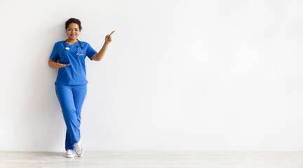 African American female nurse in blue scrubs with stethoscope leaning on wall and pointing at copy space. Confident young healthcare professional portrait with white background