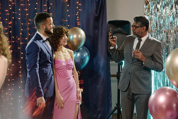 Middle aged man holding smartphone taking photo of young adult man and young adult woman standing together smiling at formal event with balloons © AnnaStills
