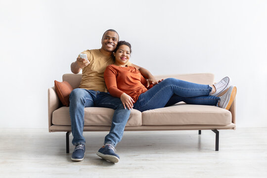 Smiling African American couple sitting on sofa with remote control, relaxing together. Portrait of young man and woman enjoying leisure time at home in casual clothing