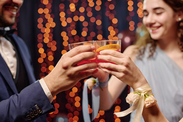 Teenagers clinking glasses during celebration, smiling and wearing formal attire, multiethnic group enjoying festive event with decorative lights in background, hands visible in foreground