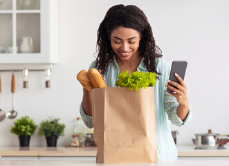 Home delivery, device, healthy food, health care and mobile app. Smiling young african american lady looks into eco package from market with vegetables and bread, holds smartphone in kitchen interior