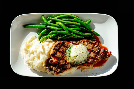 Grilled NY strip steak with green beans and mashed potatoes on a white plate against a black background
