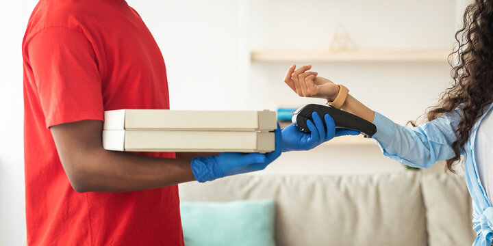 Fast Food Delivery. Smiling female customer in medical surgical mask paying using smart watch, looking at camera, black male courier wearing red uniform holding Point of Sale terminal and pizza boxes
