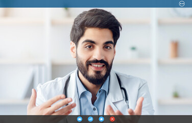 Covid-19 quarantine and medical consultation with therapist about health care. Friendly millennial arabian male doctor in white coat at table with laptop, gestures and explains in office interior