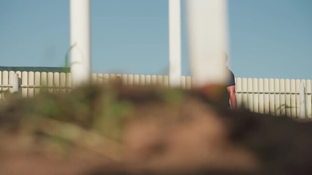 side view of farmer walking with wooden crate through white frame near green crop rows on sunny day inspecting plants for growth and health while carrying gloves and tools in belt
