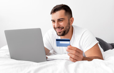 Cheerful relaxed young caucasian muscle man lies on white bed, looks at computer, holds credit card, ordering purchases, paying money in bedroom interior. Online shopping and shopaholic due covid-19