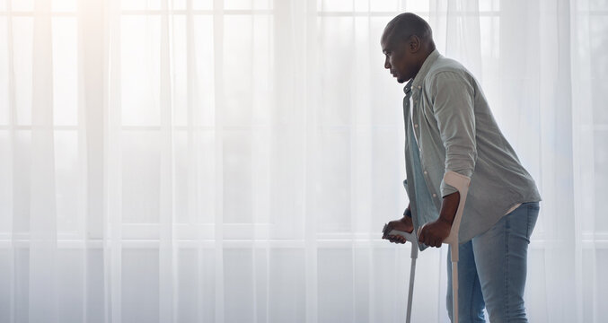Sports, physiotherapy and patient with leg injury during crutches training. Black mature disabled man stands up with crutches from wheelchair in living room interior on window with curtain background