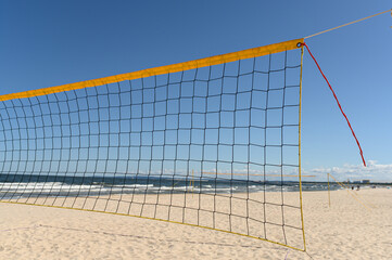 Fototapeta premium A vibrant beach volleyball net stands tall on warm sand, facing gentle ocean waves under a clear blue sky