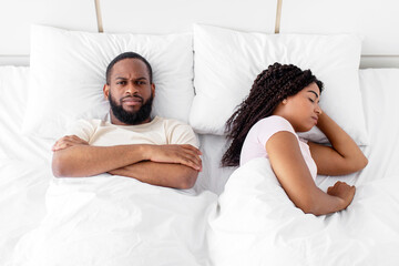 Offended millennial black male with crossed arms on his chest not sleeping and suffers from noise, angry at wife on bed in bedroom. Snoring, apnea and insomnia, problems at home during covid-19